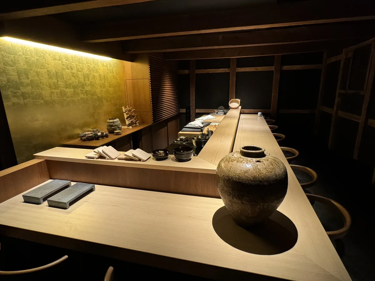金色の壁と木の梁がある落ち着いた和食店の店内。白木のL字カウンターに大きな壺が置かれている