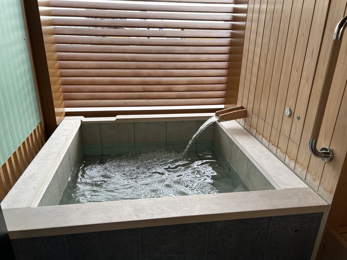 木製のルーバー窓と壁に囲まれた、四角い石造りの半露天風呂に湯が注がれている様子